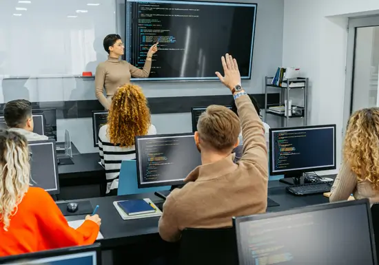 Salle de classe avec ordinateurs et écran interactif, un enseignant présente du code – solutions informatiques Aratice pour l’éducation.